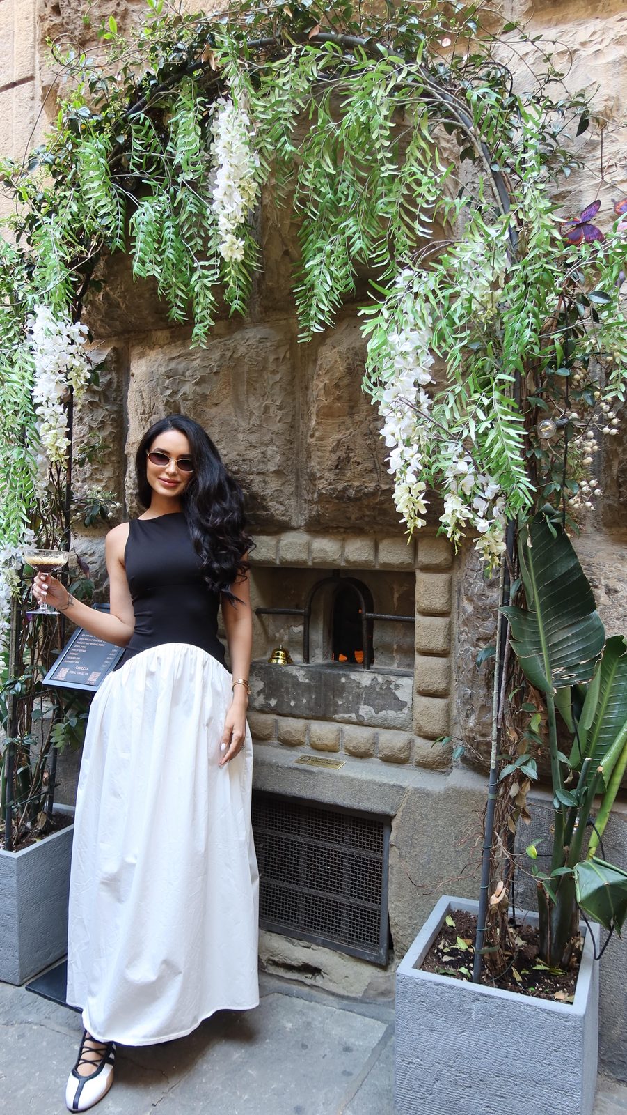 Jamillette in a black top and white skirt beneath hanging wisteria by an old stone wall in Florence