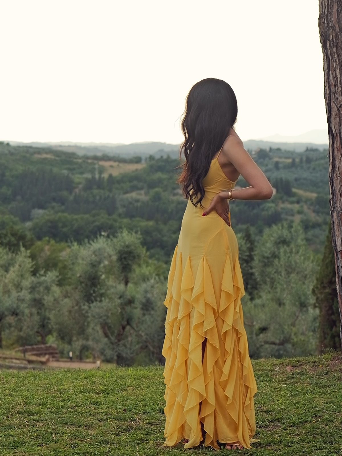 Jamillette from behind in a ruffled yellow gown, looking over green Tuscan hills