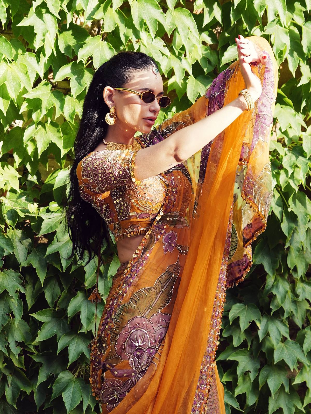 Jamillette in a saffron and violet embroidered saree, lifting the drape against an ivy wall