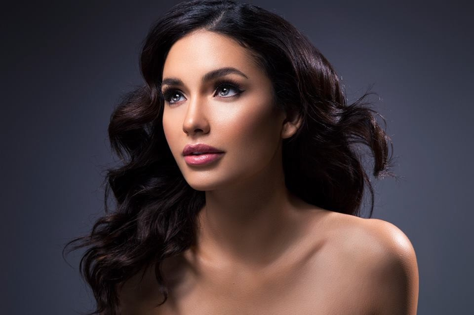 Close-up beauty portrait of Jamillette Gaxiola with long dark wavy hair and bare shoulders, looking up to the left against a dark grey-blue backdrop.