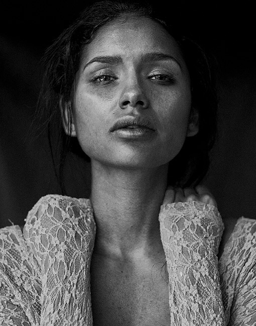 Black and white close portrait of Jamillette Gaxiola with dark hair pulled back in loose strands, head tilted up, looking down at the camera, wearing an open lace top.