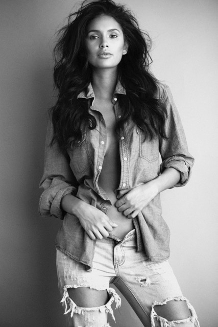 Black and white photograph of Jamillette Gaxiola with long tousled hair standing against a plain wall in an open denim shirt and ripped jeans.