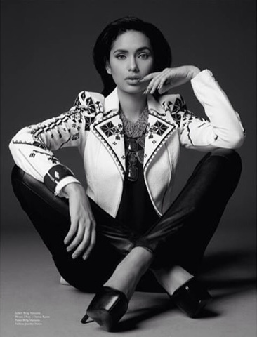 Black and white studio portrait of Jamillette Gaxiola sitting on the floor with ankles crossed, wearing an embroidered white jacket, glossy dark trousers and heels, hand at her chin.