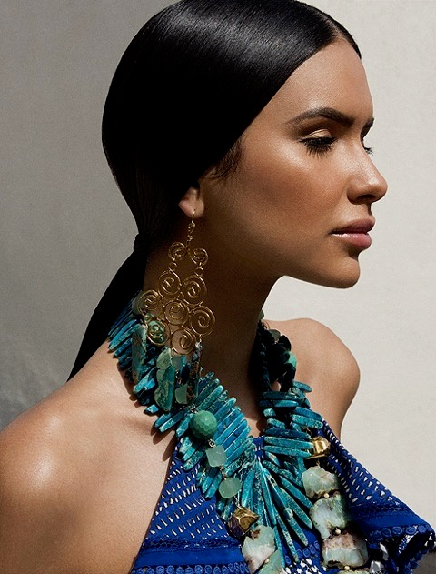 Profile portrait of Jamillette Gaxiola with sleek dark hair in a low ponytail, wearing a long gold spiral earring, a layered turquoise stone necklace and a blue halter top.