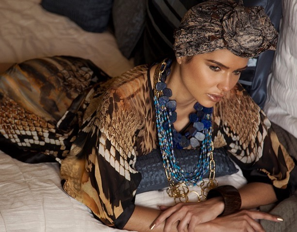 Jamillette Gaxiola in a patterned head wrap and animal-print kaftan reclines among pillows on a bed, wearing layered blue bead necklaces, gold chains and a wide dark bangle.