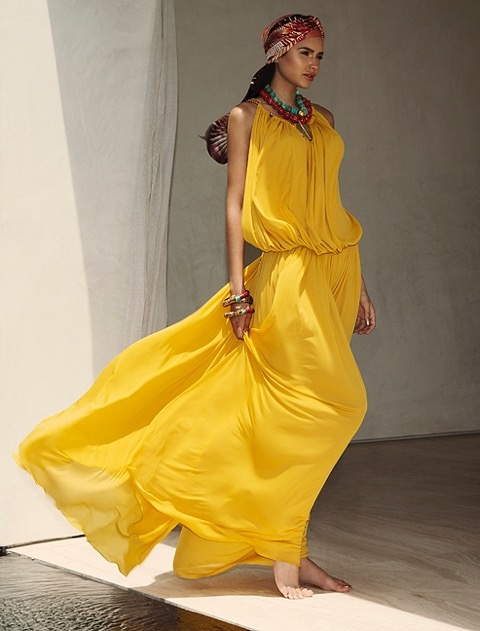 Jamillette Gaxiola in a flowing yellow halter-neck outfit and patterned head wrap walks barefoot past a pale wall, fabric billowing behind her, with chunky red and turquoise necklaces.