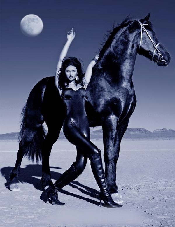 Blue-toned photograph of Jamillette Gaxiola in a glossy dark catsuit and tall heeled boots, one arm raised, standing beside a dark horse on flat dry ground under a full moon.
