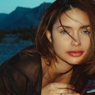 Close portrait of Jamillette Gaxiola with wind-blown brown hair across her face, leaning forward in a sheer black top, blue-toned mountains and sky blurred behind her.