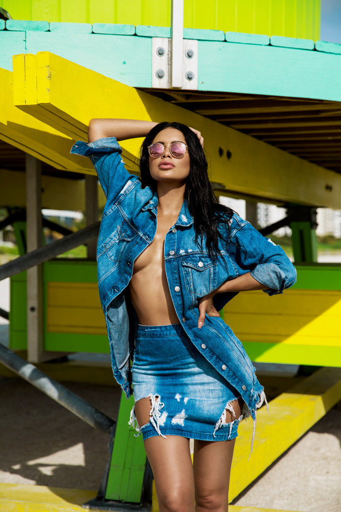 Jamillette Gaxiola in round pink mirrored sunglasses poses with one arm raised, wearing an open denim jacket and distressed denim skirt beneath a brightly painted yellow and turquoise wooden structure.